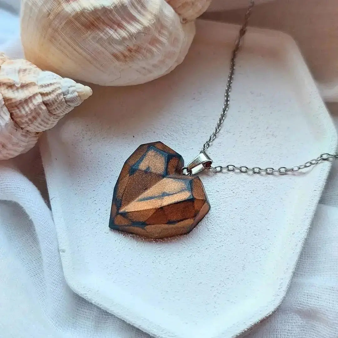 Ručno rađen privezak na lančiću u obliku srca na beloj tacni i školjkama u pozadini-slikano od gore.