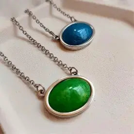 Dve ogrlice, plava i zelena, sa ovalnim priveskom na bež podlozi.